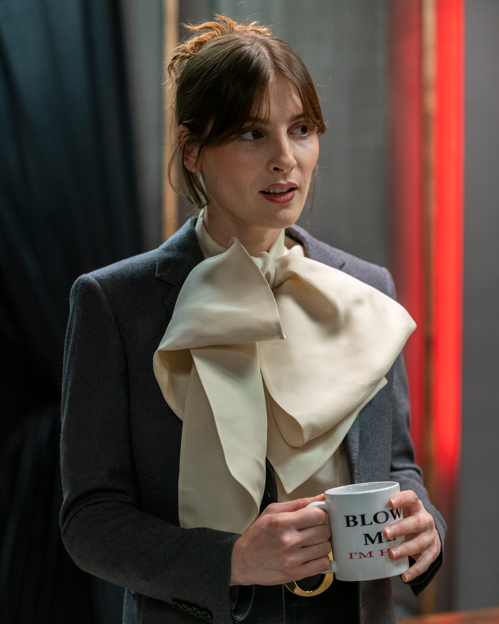 Woman wearing a blazer and oversized bow with a mug that reads 'blow me'.